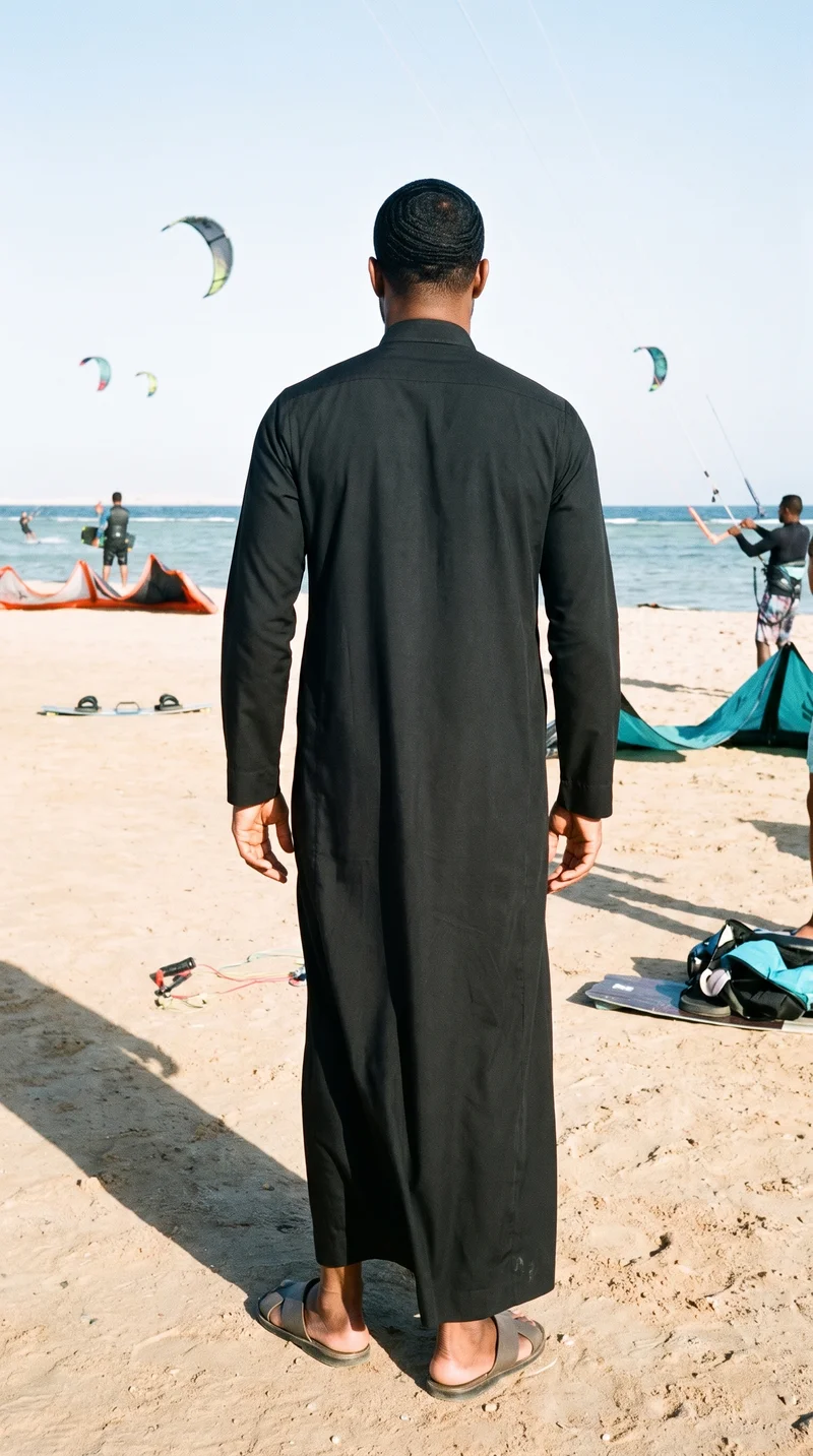 A 29-year-old African man stands in a relaxed pose, shown from the back in a full-body view wearing a black thobe. A kitesurfing beach serves as the backdrop.