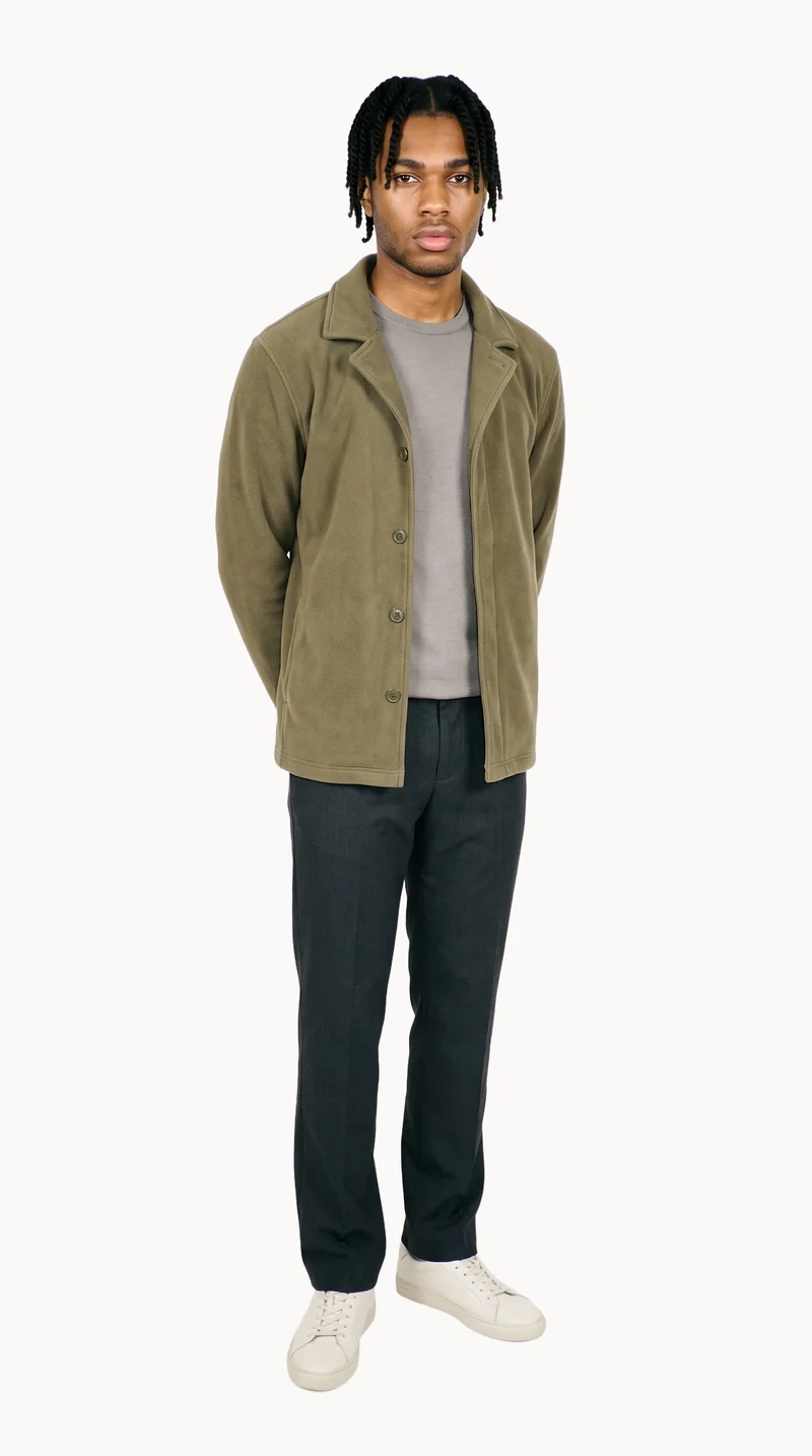 A 19-year-old African man wears an olive fleece jacket in a full-body front three-quarter view, standing with his hands behind his back and a neutral expression. A Chantilly Lace flat-color backdrop surrounds him.