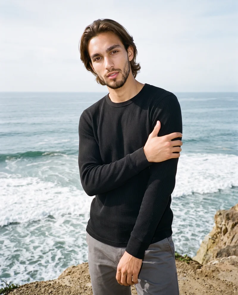 A 22-year-old Hispanic man is shown at three-quarter length from a front three-quarter angle, wearing a men's black thermal top and holding one arm. The background depicts coastal cliffs and Pacific waves.
