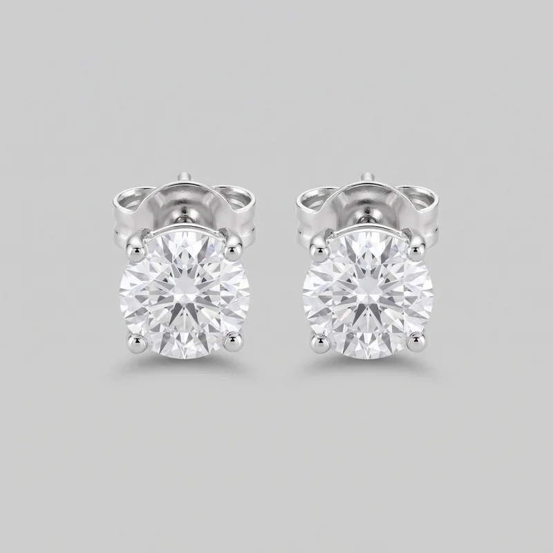 A matched pair of solitaire stud earrings for pierced ears is shown in a standalone close view. Each earring has a round faceted central stone in a four-prong setting with rounded bead-like prong tips, a curved upper basket behind the stone, a straight post, and a butterfly clutch backing visible at the rear.