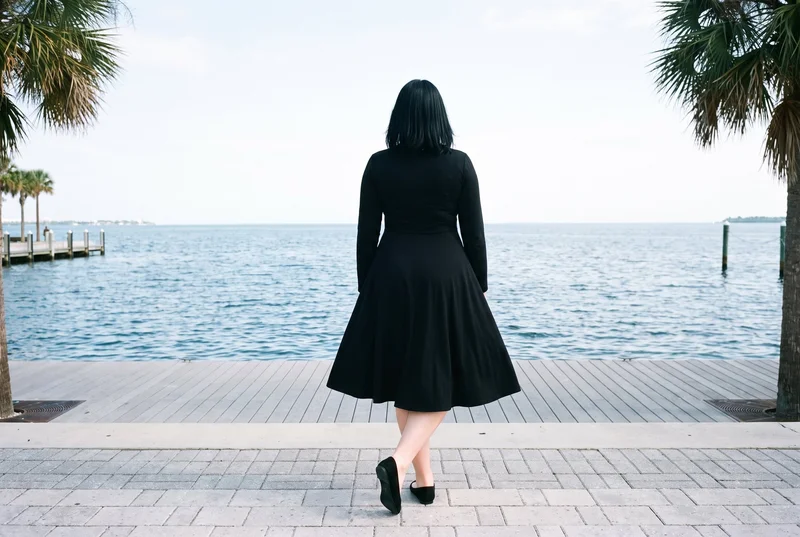 A 29-year-old Slavic woman wears a black modest maxi dress in a full-body rear view, standing with her ankles crossed. Palm trees line a waterfront promenade backdrop.