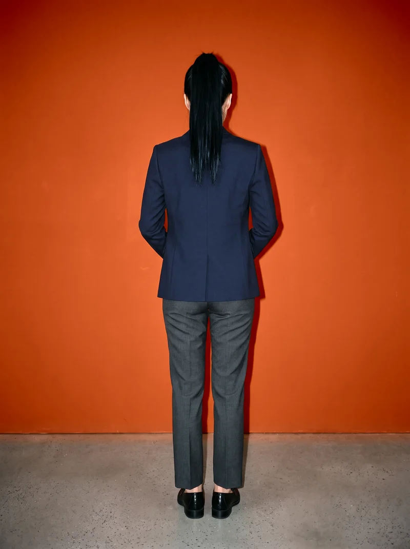 A 34-year-old Korean woman models a women's navy wool blazer in a full-body back view, standing with her hands behind her back. A rust studio-wall backdrop frames the garment.