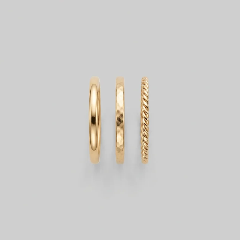Three gold ring bands stand upright side by side as a stacking ring set. The set includes a smooth domed band with rounded edges, a flat-faced band with an irregular hammered texture, and a rope-twist band with continuous diagonal raised ridges around its exterior.