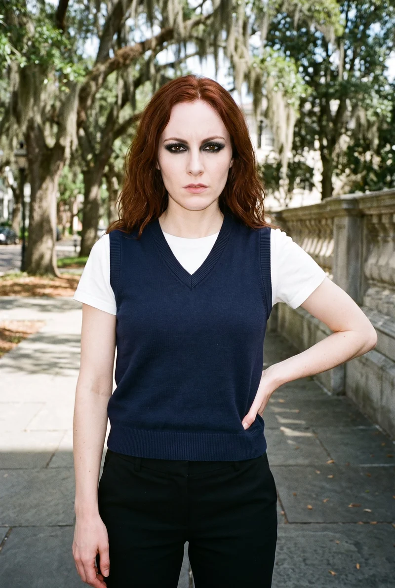 A 25-year-old Caucasian woman is shown from the upper body in a front view wearing a navy knit sweater vest. She poses with one hand on her hip and an intense facial expression, against a Spanish Moss Historic Square backdrop.