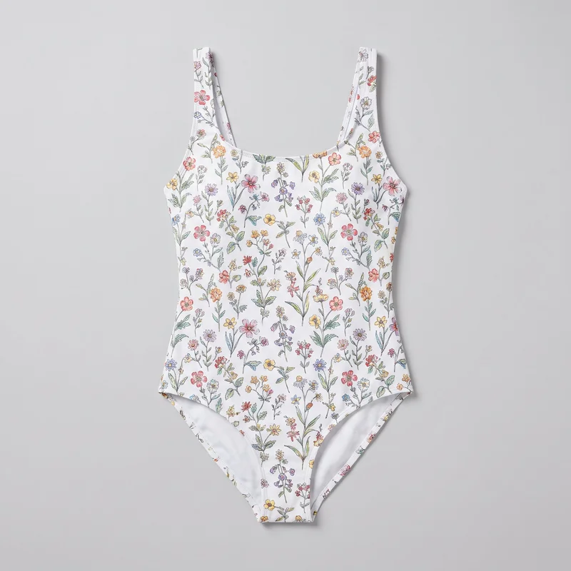 A women's one-piece swimsuit is shown in a full front flat-lay view. It has a close body-hugging fit, tank-style silhouette, wide fixed shoulder straps, a rounded scoop neckline, low-cut arm openings, high-cut leg openings, and a tapered brief-style bottom. Bound edges outline the neckline, straps, armholes, and leg openings, with side seams and a visible center seam through the lower front. Scattered stems, leaves, and blossoms form an all-over fine botanical floral print across the exterior.