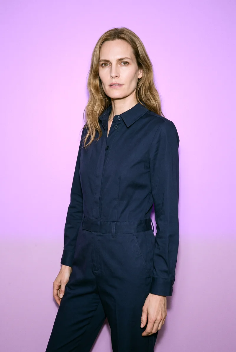 A 51-year-old Caucasian woman wears a navy tailored jumpsuit in a three-quarter-length view. She stands in a relaxed pose at a front three-quarter angle with a neutral facial expression, against a mauve studio-wall backdrop.