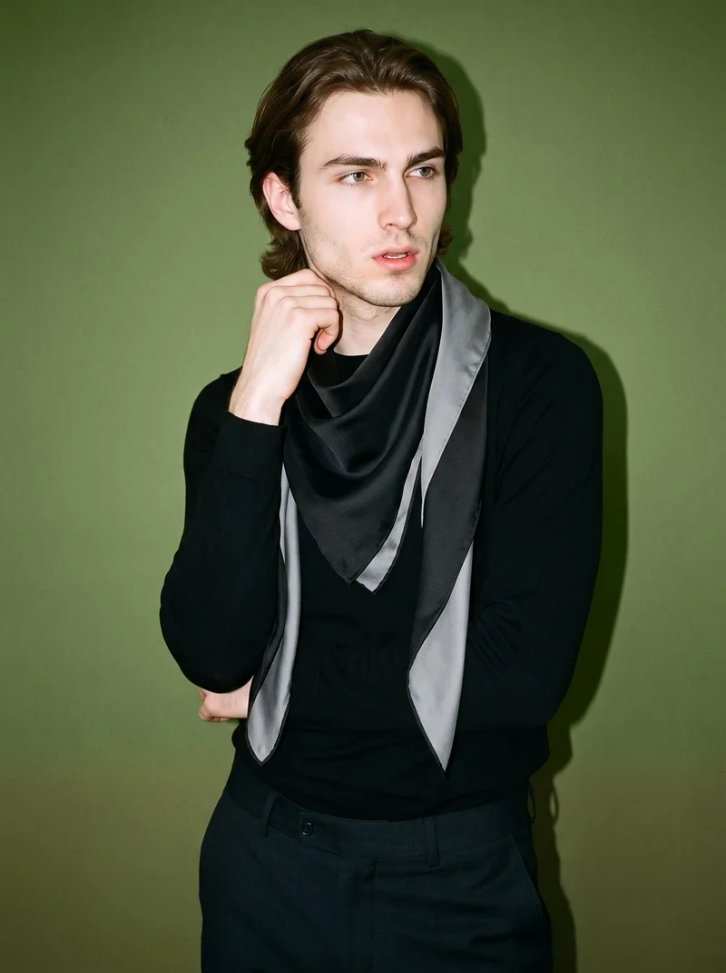 A 22-year-old Caucasian man is shown in an upper-body, angled-front view wearing a men's black silk scarf. His wrist is held at his jaw in an editorial pose, with an olive grove backdrop.