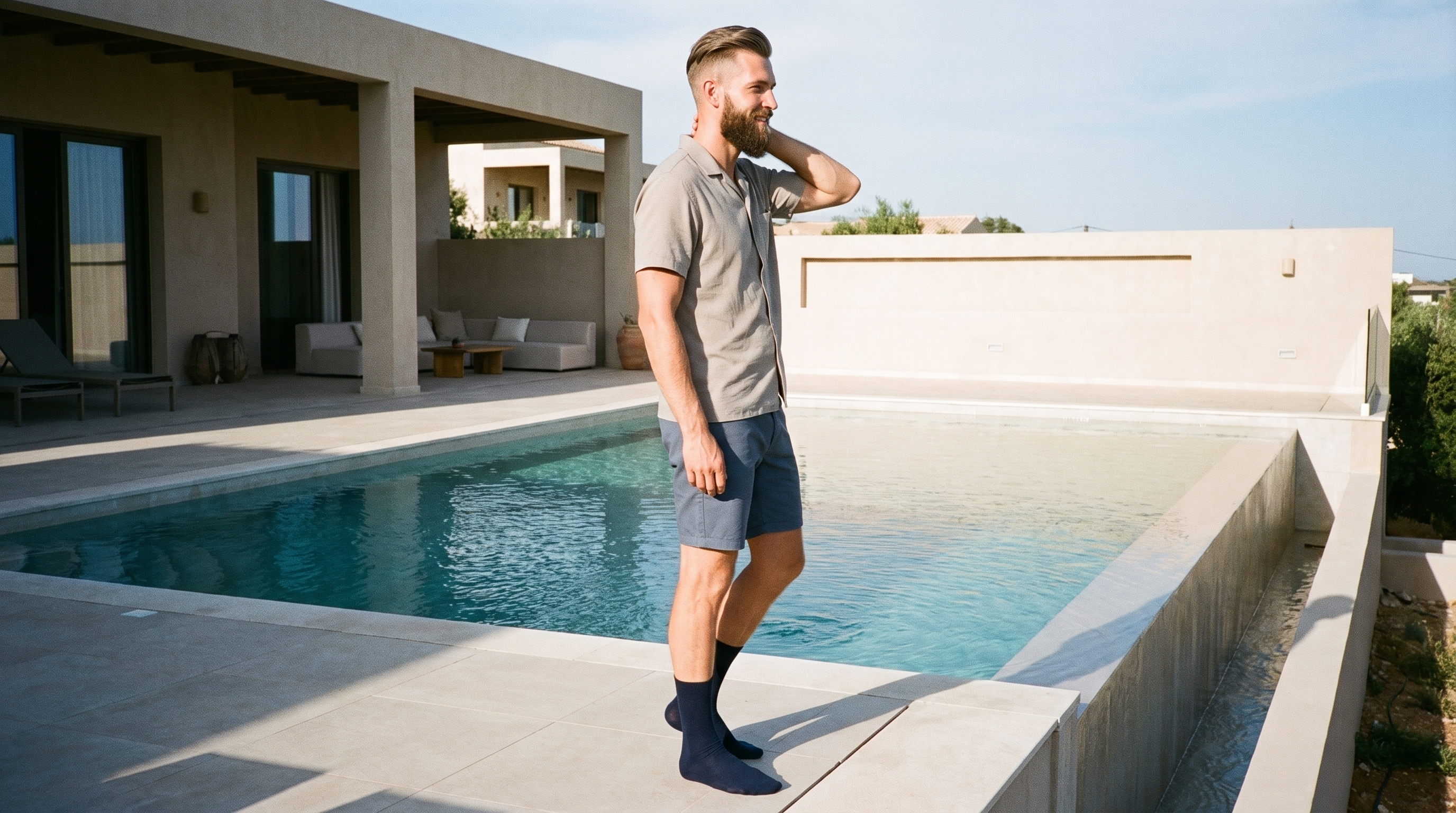 A 33-year-old Caucasian man is shown in side view wearing men's navy dress socks, with one hand behind his neck and a smile. A villa infinity pool forms the environmental backdrop.