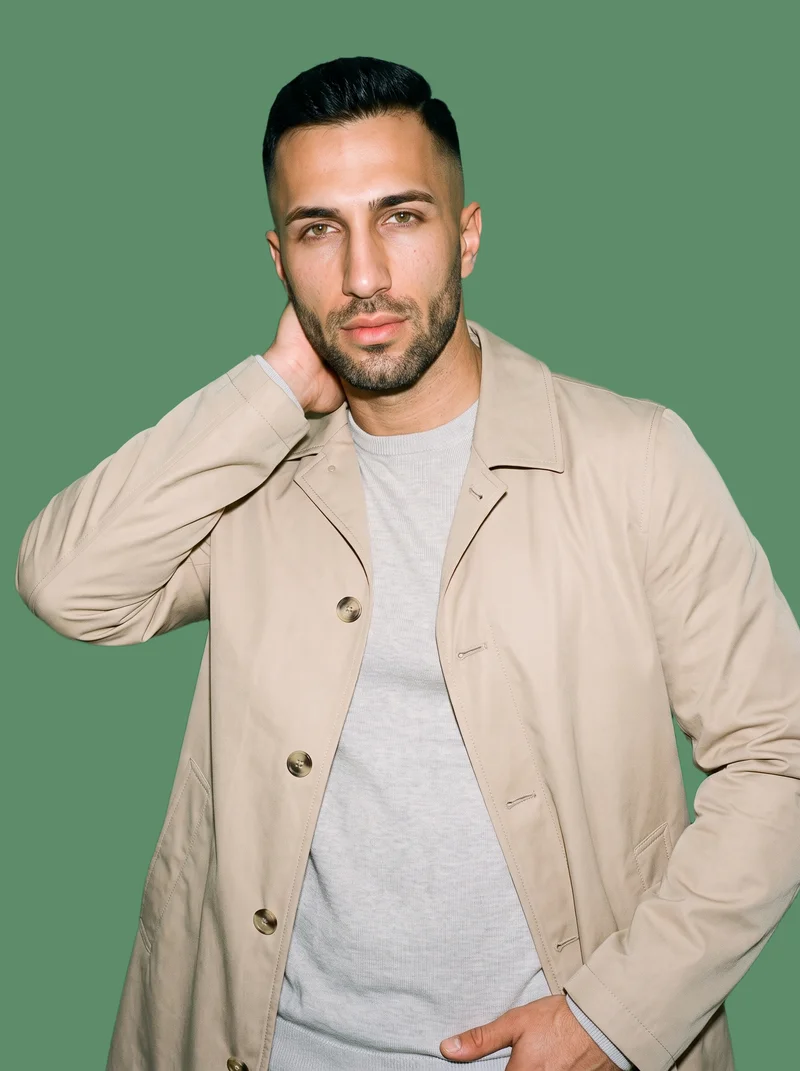 A 35-year-old Turkish man is shown from the upper body at a front three-quarter angle, wearing a beige cotton mac with one hand behind his neck. The eucalyptus-colored flat backdrop frames the studio-cutout composition.