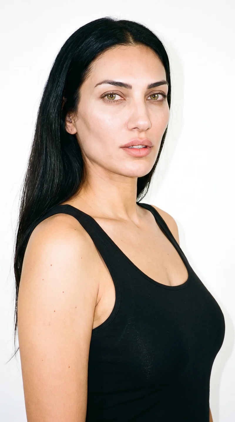 This 40-year-old Turkish woman has a medium skin tone with a neutral undertone. Her face is long, with more length than width, a tall forehead, and elongated cheeks. She has medium-thickness eyebrows, hazel eyes with green-gold flecks, and full lips. Her long, deep black hair is worn loose past the shoulders with natural volume.