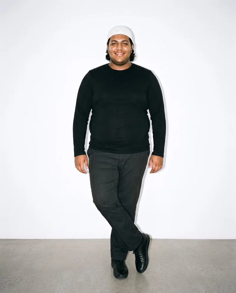 A 22-year-old Arab man wears a white knit kufi in a full-body front view, standing with his ankles crossed and a natural expression. A porcelain studio-wall backdrop fills the background.