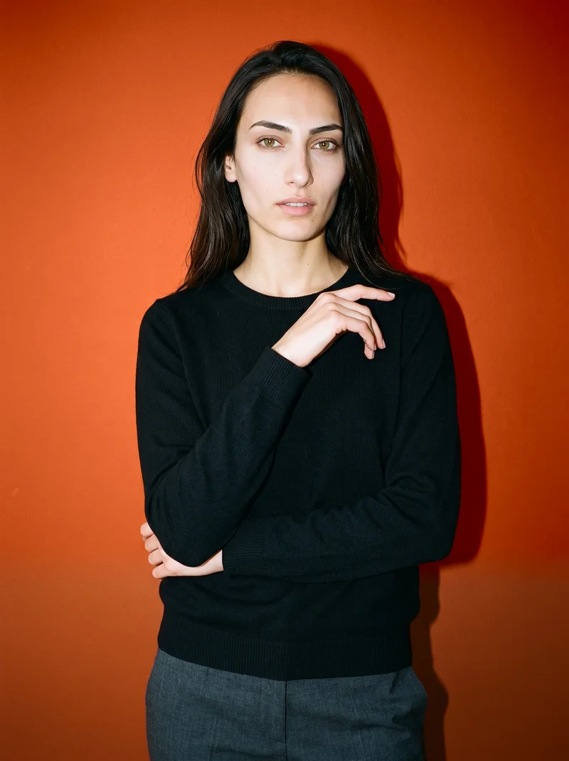 A 20-year-old Turkish woman is shown from the front in an upper-body view wearing a black pullover jumper. She is touching her wrist and has a soft facial expression, with a rust studio-wall backdrop.