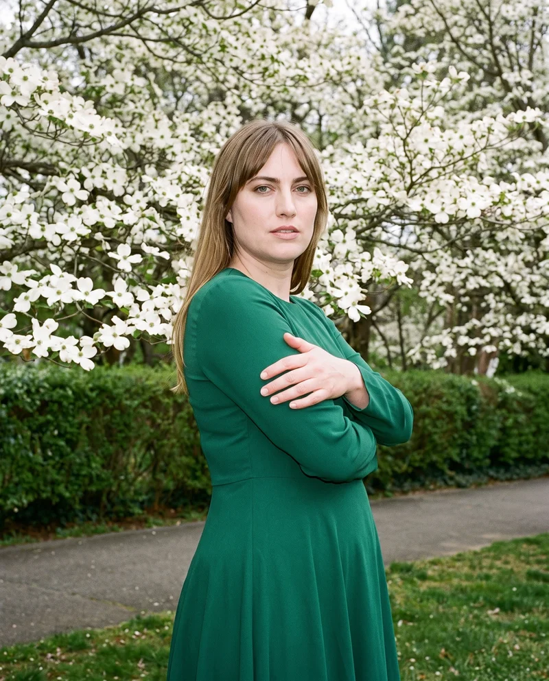 A 41-year-old Caucasian woman wears an emerald green modest dress in a three-quarter-length view from a front three-quarter angle. She stands with her arms crossed and a confident expression, with a dogwood tree grove serving as the backdrop.