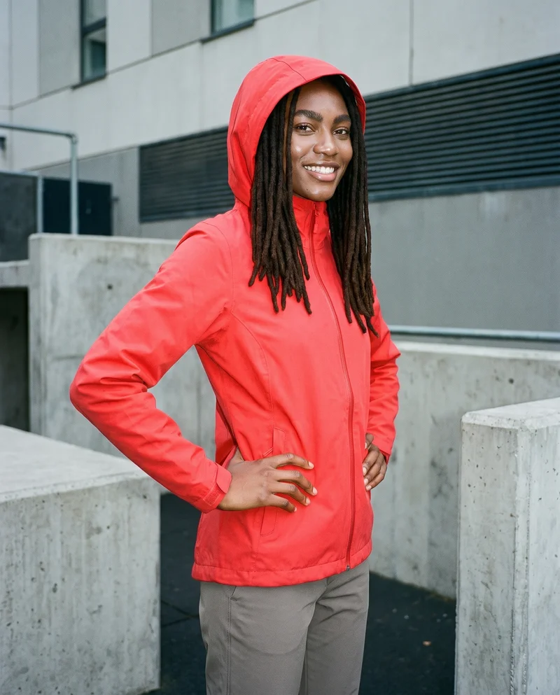 A 19-year-old African woman wears a red hardshell jacket in a three-quarter-length composition. She is shown from a front three-quarter angle with her hands on her waist and a smile, against a parkour urban obstacles backdrop.