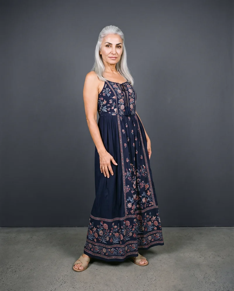A 58-year-old Turkish woman models a navy boho maxi dress in a front-view environmental composition, posing with a waist twist and a curious expression. A graphite studio-wall backdrop frames the scene.