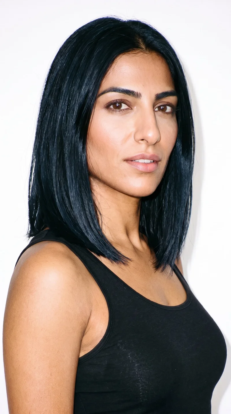 A 34-year-old Arab woman with caramel, neutral-undertone skin and an inverted-triangle face shape, defined by a wider forehead that narrows toward the chin. She has thick eyebrows, medium brown eyes, and medium-sized lips. Her deep black hair is styled in a long bob that falls between the chin and shoulders.