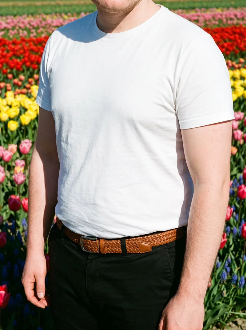 A male model is shown from the torso at an angled front view, wearing a tan woven leather belt with his arms relaxed. A tulip field forms the backdrop.