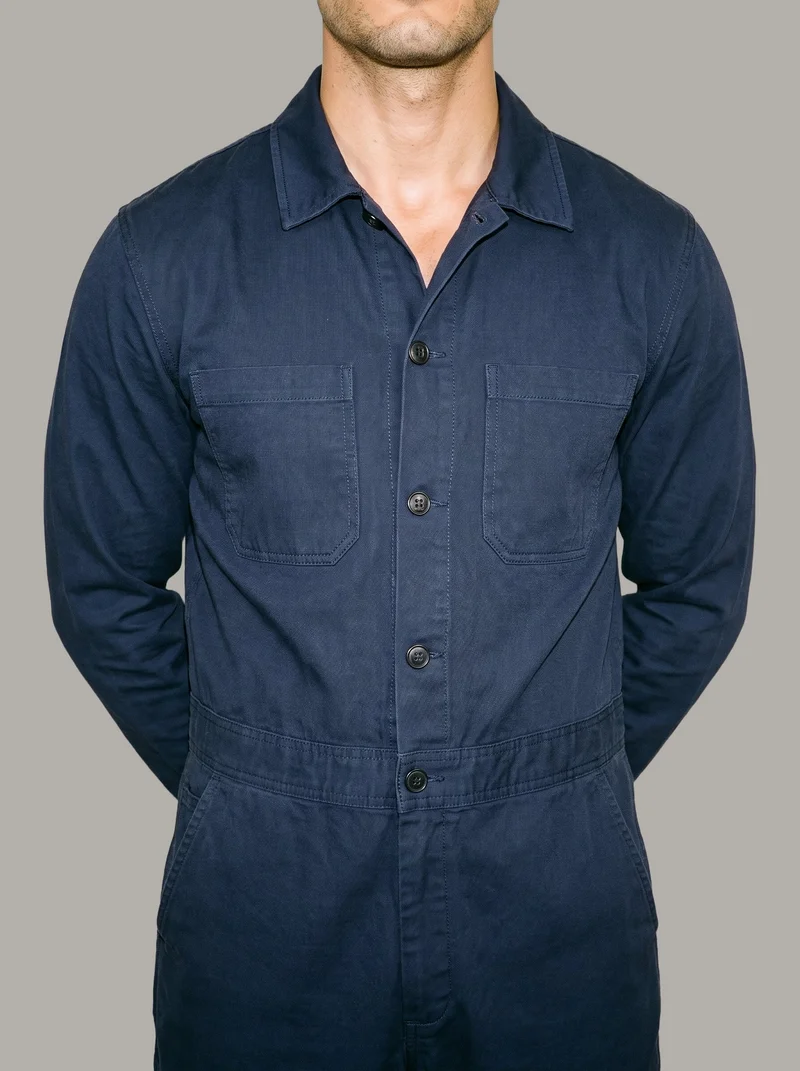 A man wears a navy cotton boilersuit, shown from the front in a torso crop with his hands behind his back. A flat Couture Grey backdrop surrounds the studio-cutout composition.