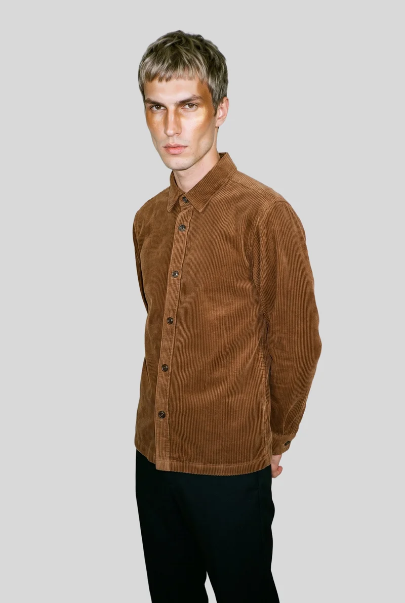 A 27-year-old Slavic male model wears a brown corduroy overshirt in a three-quarter-length, angled front view with his hands behind his back. The model is shown against a flat pearl grey backdrop.