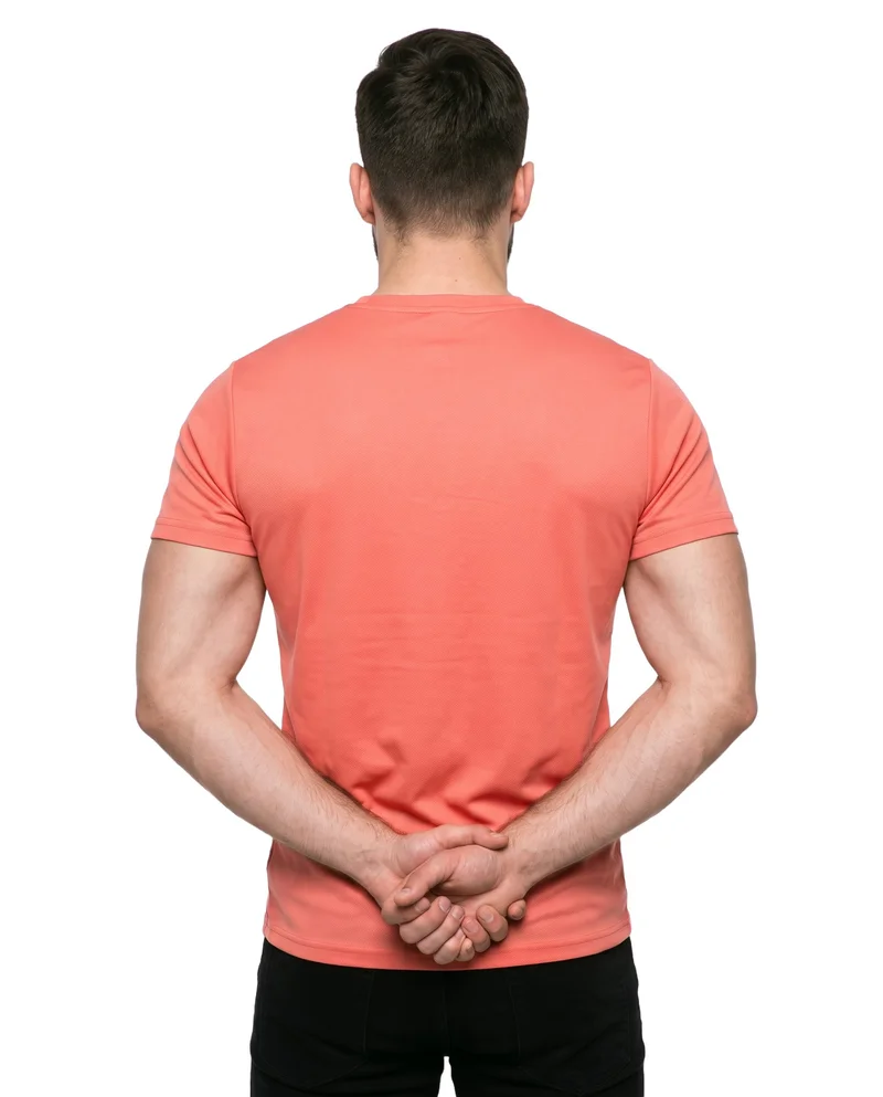 A 22-year-old Slavic male model is shown from the rear in an upper-body view, wearing a coral seamless performance top with his hands behind his back. The garment is presented against a pure white backdrop.