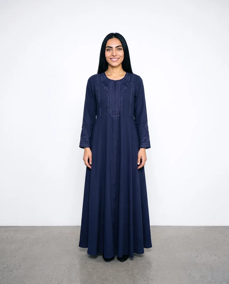A 22-year-old Arab woman stands straight in a full-body front view wearing a navy embroidered abaya. Her natural expression is visible against a porcelain studio-wall backdrop.