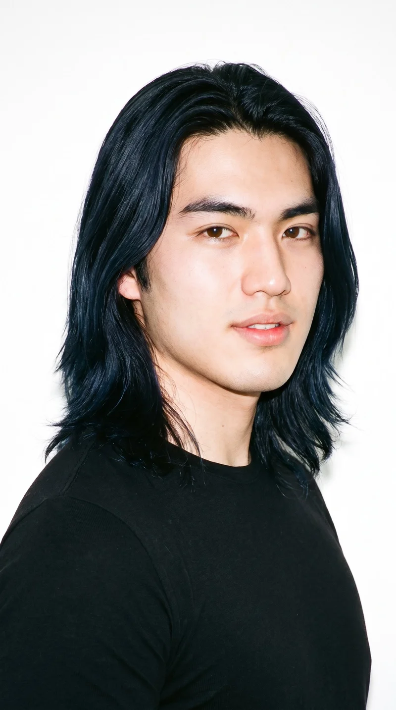 A 19-year-old Japanese man with East Asian facial features and light porcelain skin with a warm yellow undertone. His face has balanced oval proportions and a softly rounded jaw, with thick eyebrows, brown eyes, and full lips. Long, deep black hair falls loose past his shoulders with natural volume. He is clean-shaven, with smooth skin across his face.