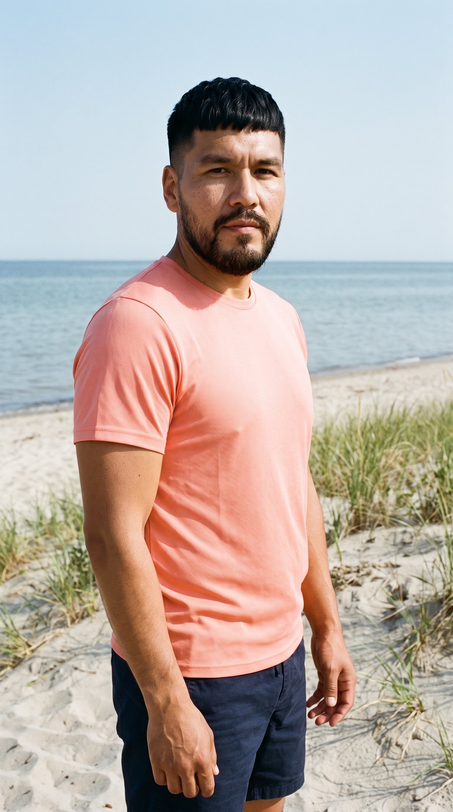 A 38-year-old Indigenous American man wears a coral seamless performance top in an upper-body, angled-front view with his arms relaxed and a confident expression. The Martha's Vineyard Beach backdrop is visible behind him.