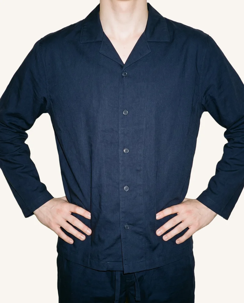 A male model is shown from the torso in a front-facing pose with his hands on his hips, wearing a navy cotton pyjama set. The garment is presented against a soft ivory backdrop.