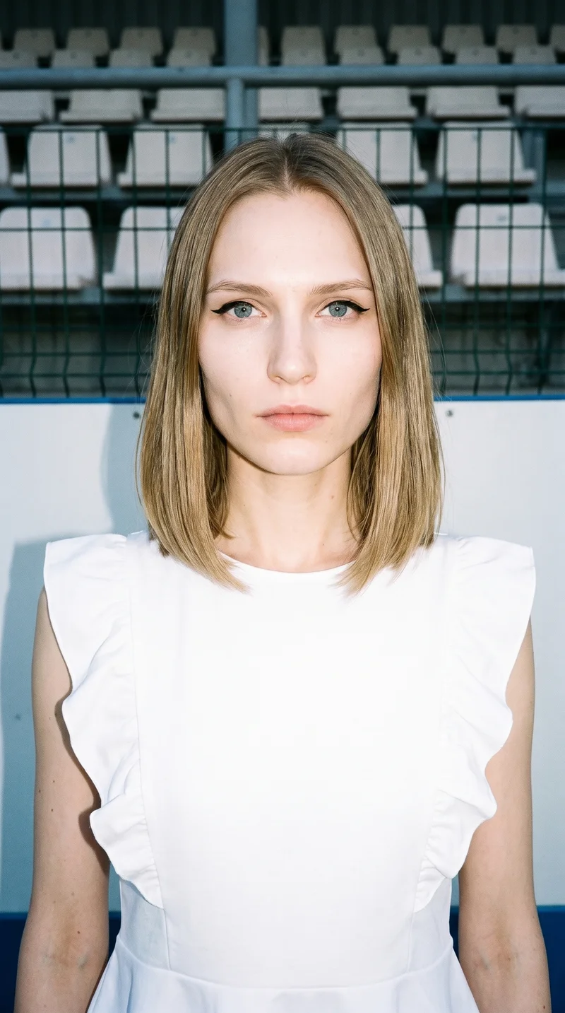 A 27-year-old Slavic woman models a white peplum top in a front-facing bust view with her shoulders square and a neutral expression. Stadium seating forms the backdrop.