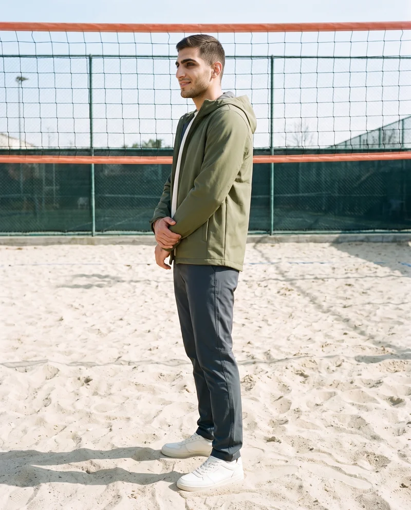 A 22-year-old Arab man is shown full body in side view, smiling with his arms held low while wearing a green waterproof jacket. A beach volleyball court forms the backdrop.
