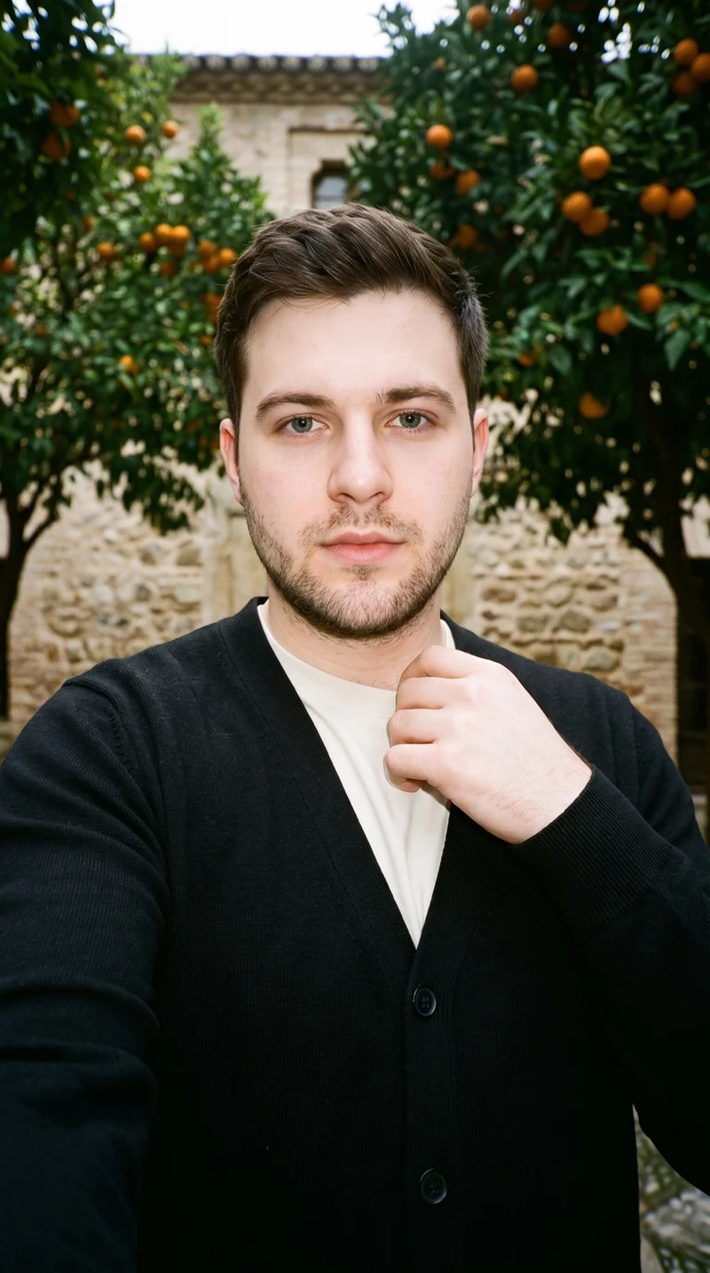 A 26-year-old Caucasian man wears a black button-up cardigan in a front-facing selfie, with one hand at his collarbone and a neutral expression. A Seville courtyard forms the backdrop.