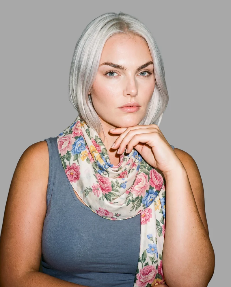 A 23-year-old Caucasian woman wears a floral print shawl in a bust-framed front three-quarter view. She rests her chin on her knuckles with a curious facial expression, against a flat pebble grey backdrop.