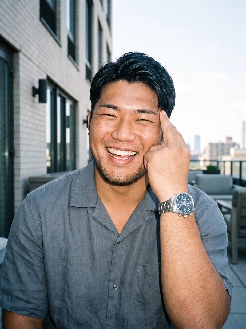 A 31-year-old Japanese man is shown from the front in an upper-body view, laughing with one hand at his temple while wearing a silver chronograph sports watch. A penthouse terrace forms the backdrop.