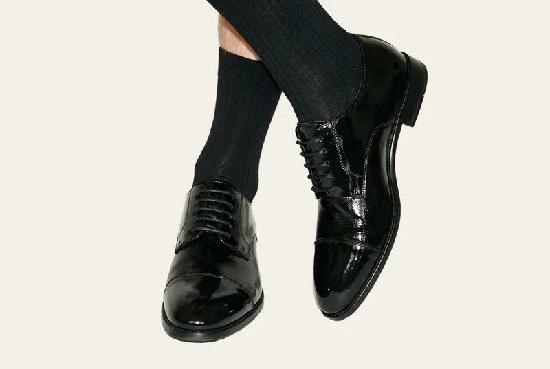 Black patent dress shoes worn on both feet, shown from a front three-quarter angle with the ankles crossed. The shoes are set against an alabaster flat-color backdrop.