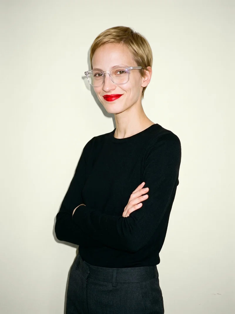 A 25-year-old Caucasian woman wears clear-frame blue-light glasses in an upper-body, angled-front view. She is smiling with her arms crossed against an ivory lace backdrop.