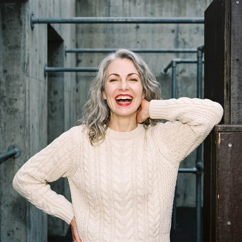 A 55-year-old Caucasian woman wears a cream cable-knit pullover jumper in an upper-body front view. She is laughing with one hand behind her neck, against a parkour urban obstacles backdrop.