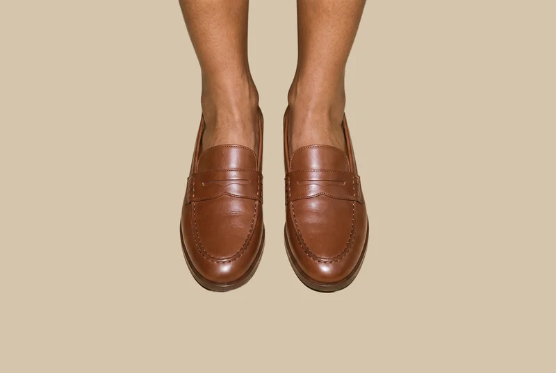A pair of women's brown penny loafers shown from the front with both feet together against a Venetian plaster-style flat-color backdrop.