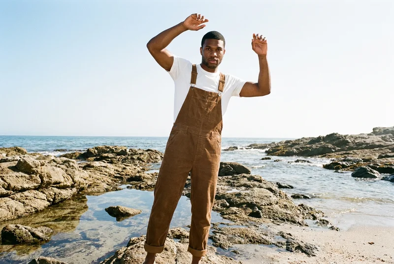 A 22-year-old African man wears men's corduroy dungarees in a front-facing environmental view, standing with his arms overhead and a natural expression. A rocky beach forms the backdrop.