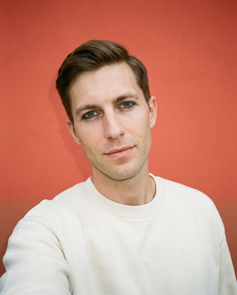 A 36-year-old Caucasian man wears a cream oversized crewneck sweatshirt in a front-facing selfie with his chin down and a smile. A terra cotta studio-wall backdrop fills the background.