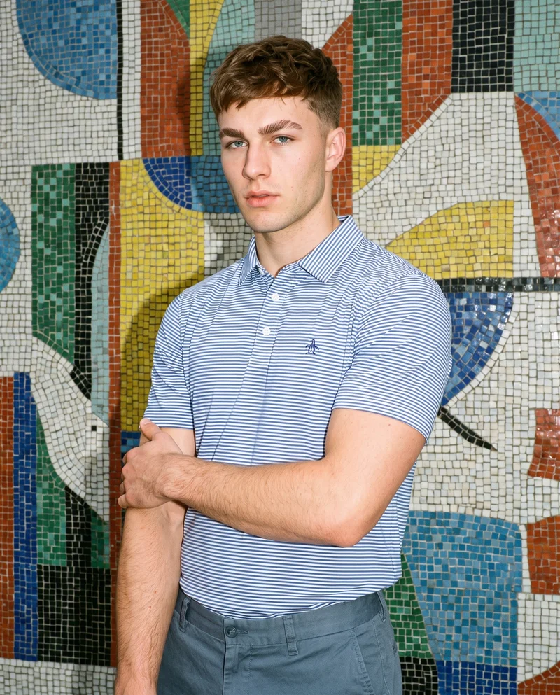A 20-year-old Caucasian male model wears a men's striped cotton polo shirt in an upper-body view from a front three-quarter angle. He holds himself in a self-hug pose with a neutral expression, with a Barcelona modernist building backdrop.