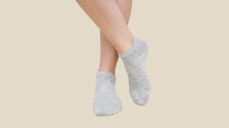 A person's feet are shown from the front with the ankles crossed, wearing women's grey cotton ankle socks. A flat cashmere-colored backdrop fills the background.