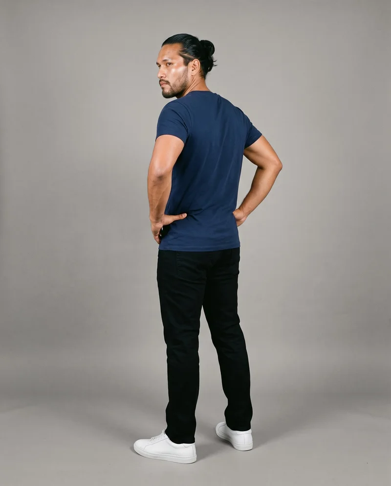 A 35-year-old Indigenous American man wears a navy V-neck T-shirt in a full-body, angled rear view with his hands on his hips. The shirt is shown against a flat Couture Grey backdrop.