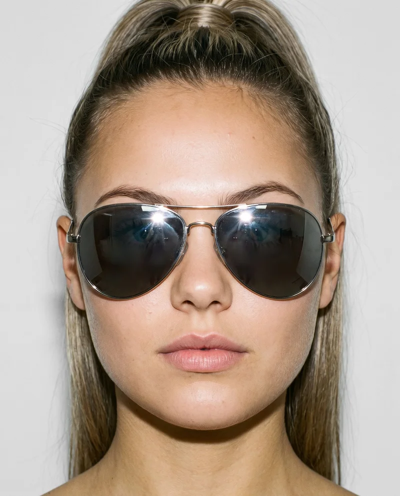 A 21-year-old Caucasian woman wears women's mirrored sunglasses in a head-only, front-view composition. Her head is held level against a cloud-white studio-wall backdrop.