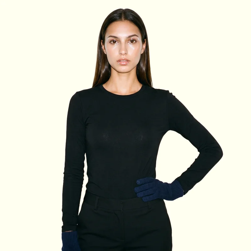 A 21-year-old Slavic woman is shown from the upper body in a front-facing pose with one hand on her hip, wearing women's navy touchscreen gloves. Her expression is neutral, and an ivory lace backdrop frames the studio-cutout composition.