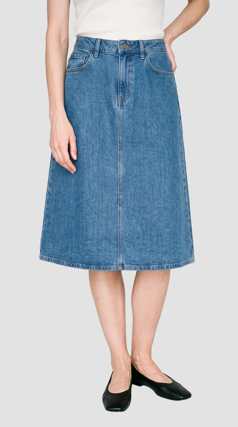 Denim A-line skirt shown from the front in a lower-body crop, with the model’s ankles crossed. The skirt is presented against a flat fog-grey backdrop.