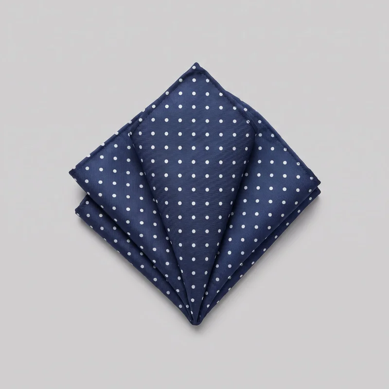 A standalone pocket square folded into a structured pointed puff arrangement, with layered triangular peaks, overlapping panels, and bordered edges visible. The square handkerchief has a fine ribbed weave texture, diagonal fold lines, and an all-over small polka-dot pattern across its visible surfaces.