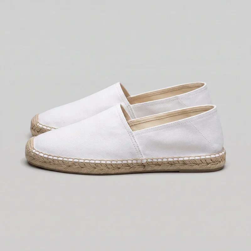 A pair of white men's slip-on espadrille shoes is shown in a side three-quarter view, with both shoes mostly visible. The shoes have rounded closed toes, low-cut oval openings, smooth plain vamps, stitched collar edges, side seam panels, and small notches at the vamp openings. Braided rope midsoles with looped perimeter stitching and thin flat outsoles complete the design; no laces, buckles, zippers, logos, prints, or embellishments are visible.