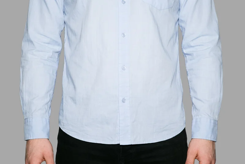 A light blue button-down shirt is shown from the front in a hip-level frame, with the model’s hips level. The shirt is presented against a flat pebble grey backdrop.